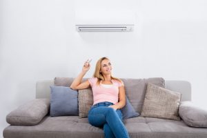 Young Happy Woman Sitting On Couch Operating Air Conditioner With Remote Control At Home